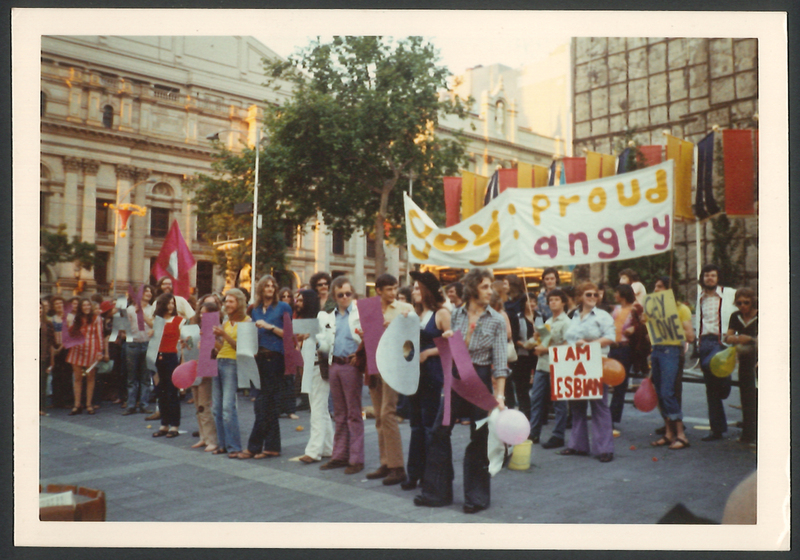 Colour photograph of a group of people with banner 'GAY: PROUD ANGRY' waiting to begin a parade.