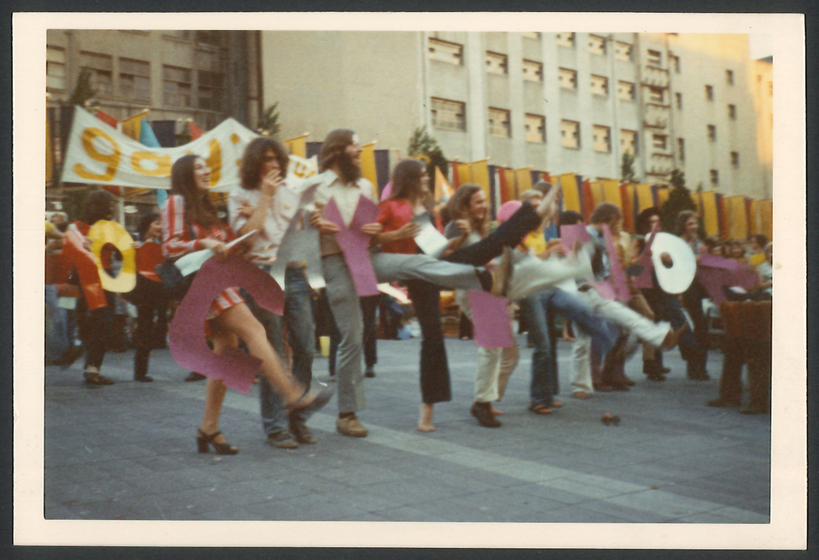 Colour photograph of a line demonstrators doing a high kick in public square with banner 'GAY'.