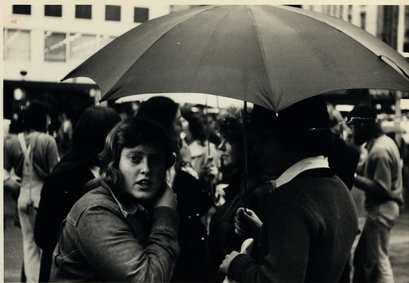 Black and white photograph of a woman huddled under an umbrella in a crowd in a city street.