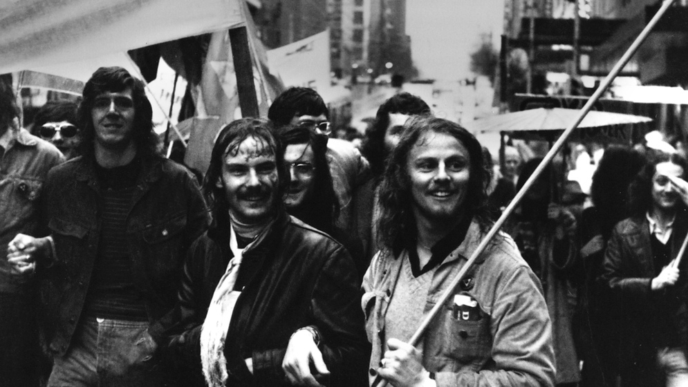 Black and white photograph of demonstrators with banners in a city street, including two men arm in arm.
