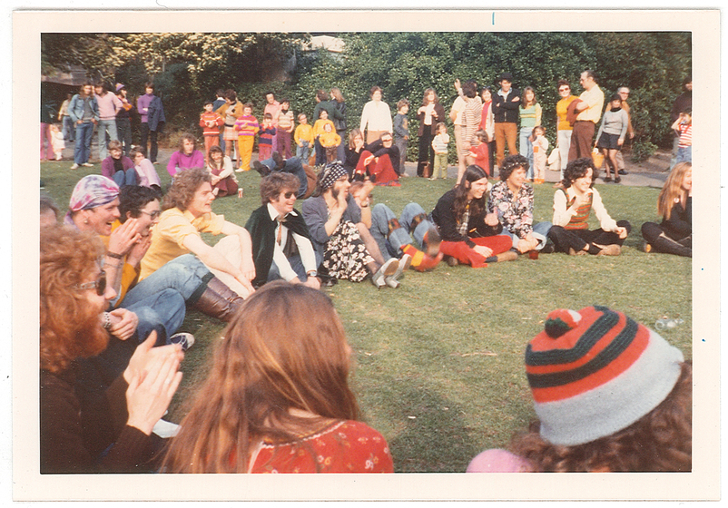 Colour photograph of about fifty people sitting on a lawn, trees behind.