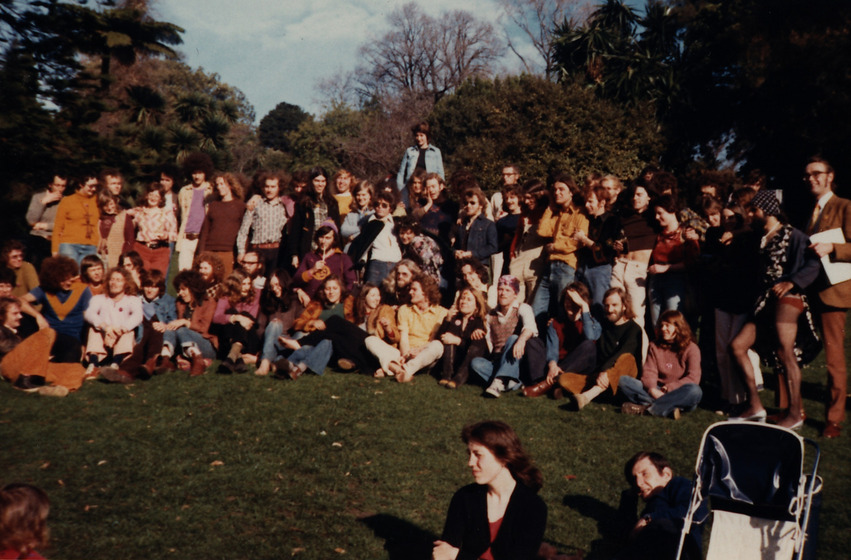 Group colour photograph of about fifty people sitting and standing on a lawn with trees behind.