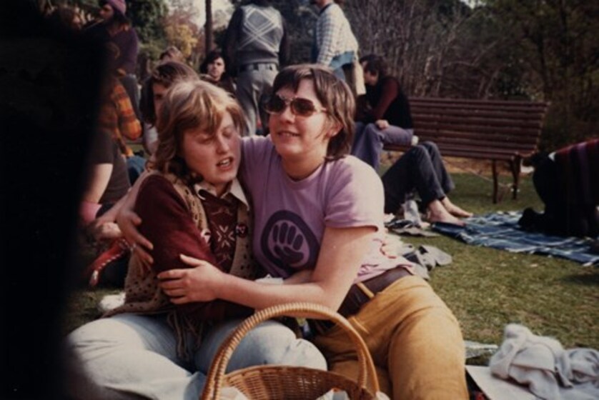 Two women embracing sitting on a lawn. Other people and trees behind them.