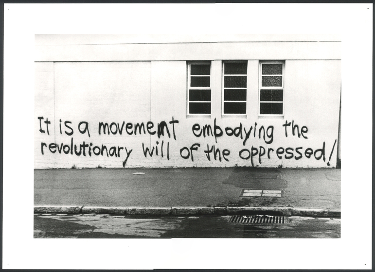 Black and white photograph of graffiti spray painted on a wall.