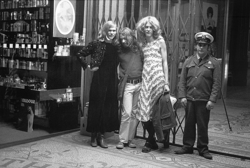 Black and white photograph of two men in drag, a male companion and a male security guard outside a shopfront.