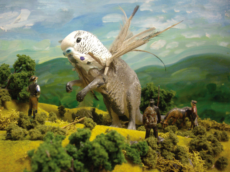 Model of budgie head on winged kangaroo body, towering over white and Aboriginal men on hill in front of painted background.