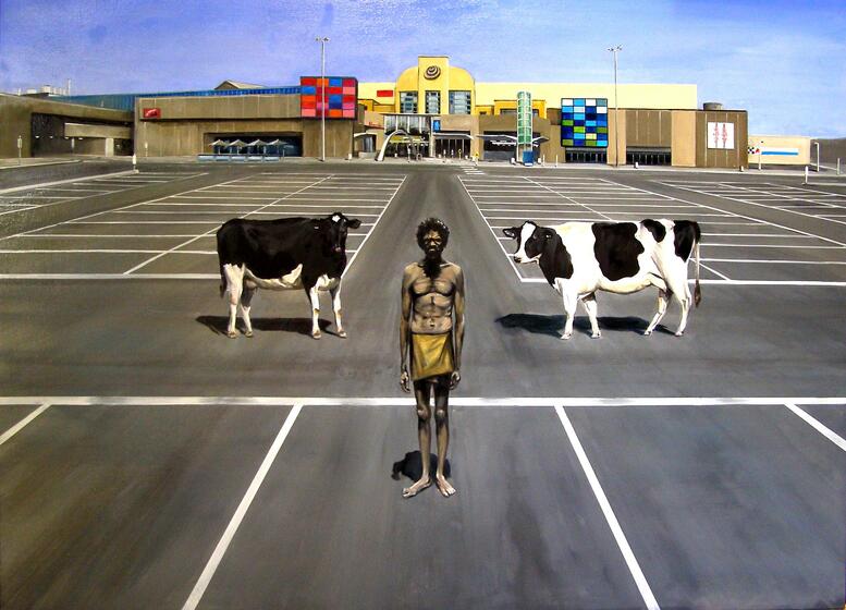 Traditionally dressed Aboriginal man standing in an empty shopping centre carpark with two cows standing behind. 