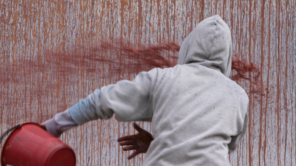 Back of person in grey hoodie covering their head, moving a red bucket toward a grey wall covered in splattered red paint.  