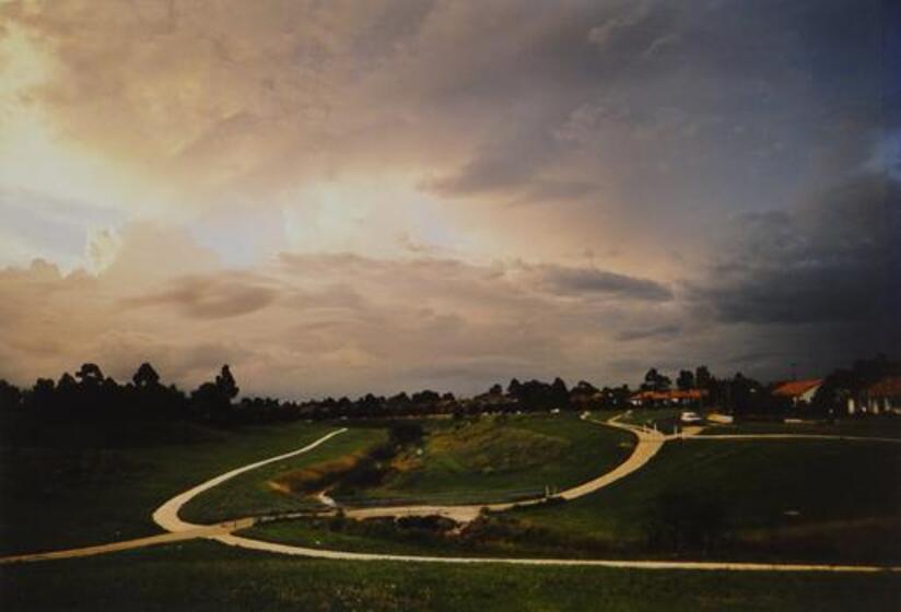Creek in grass area with footpaths intersecting near small bridge. Trees and houses in distance with dark, cloudy sky.   