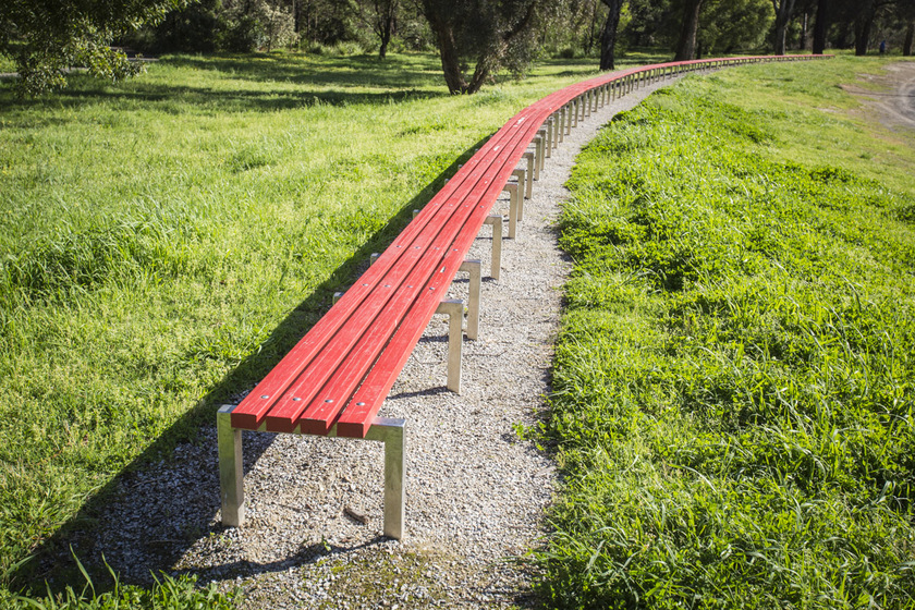 Long red wooden bench on concrete footing, curving around grassed oval with trees in background.