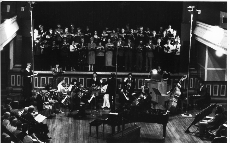 Black and white photo, inside a Town Hall. Background: A chorus of people standing on the raised stage. Centre: musicians. Foreground: Audience to left and right, piano in centre.