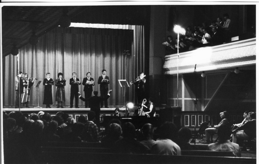 Black and white photo, inside a Town Hall. Background left: A small chorus of people standing on the raised stage with curtains closed behind. Right: A chorus of people standing in the dress circle with song sheets. Foreground: Audience in seating on the floor of the Town Hall.