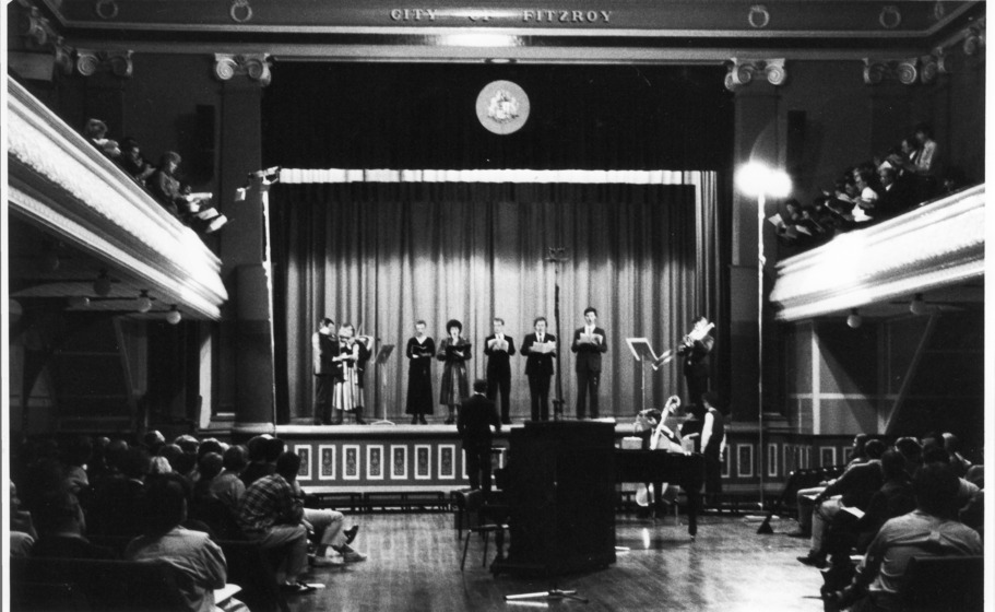 Black and white photo, inside a Town Hall. Background centre: A small chorus of people standing on the raised stage with curtains closed behind. Right and left: A chorus of people standing in the dress circle with song sheets. Middle and foreground: Audience in seating on the floor of the Town Hall left and right. Piano in centre foreground.