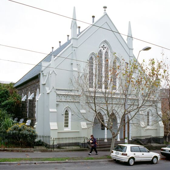 Buiding facade of pitch roofed, pale blue fronted bluestone brick church with three-arch entry and upper stained glass windows.