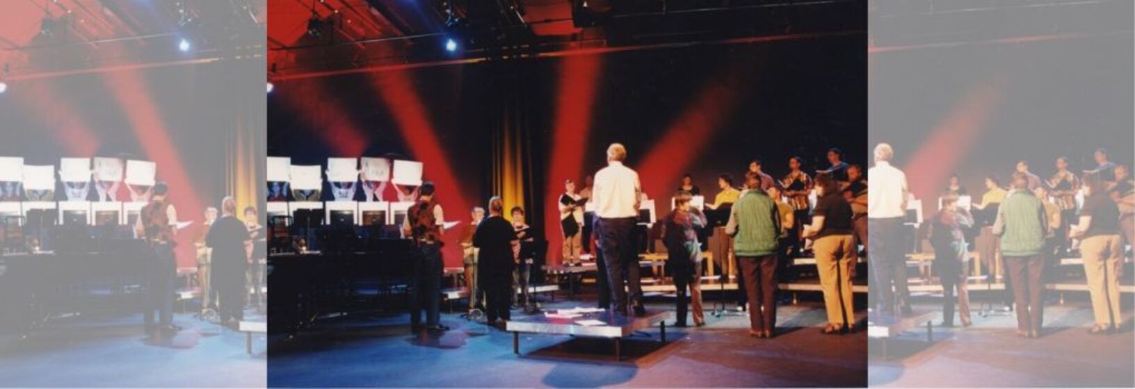 Choir of people standing on tiered platform in theatre with red lighting and curtain backdrop, conductor in centre (back to camera)