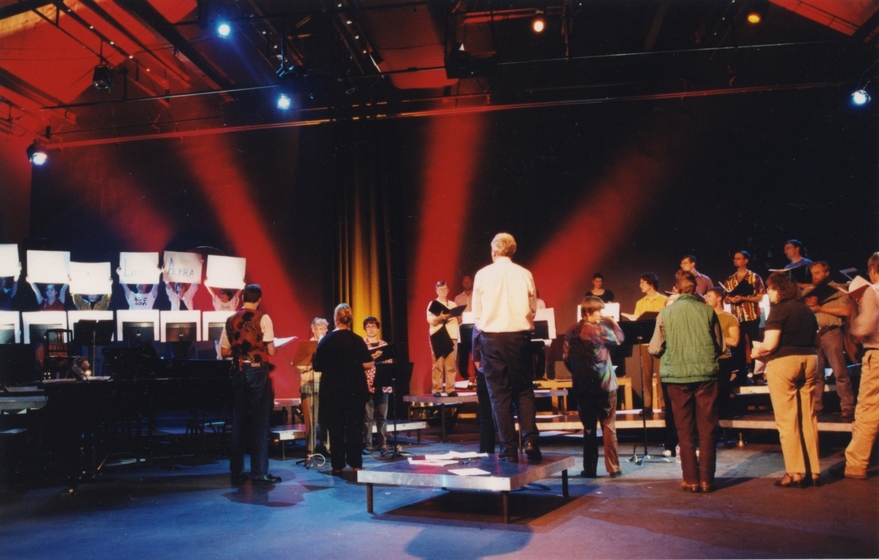 Choir of people standing on tiered platform in theatre with red lighting and curtain backdrop, conductor in centre (back to camera)