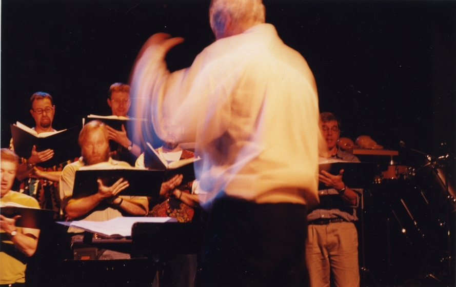 Two rows of choir singers holding music, with conductor facing them, left arm blurred with movement