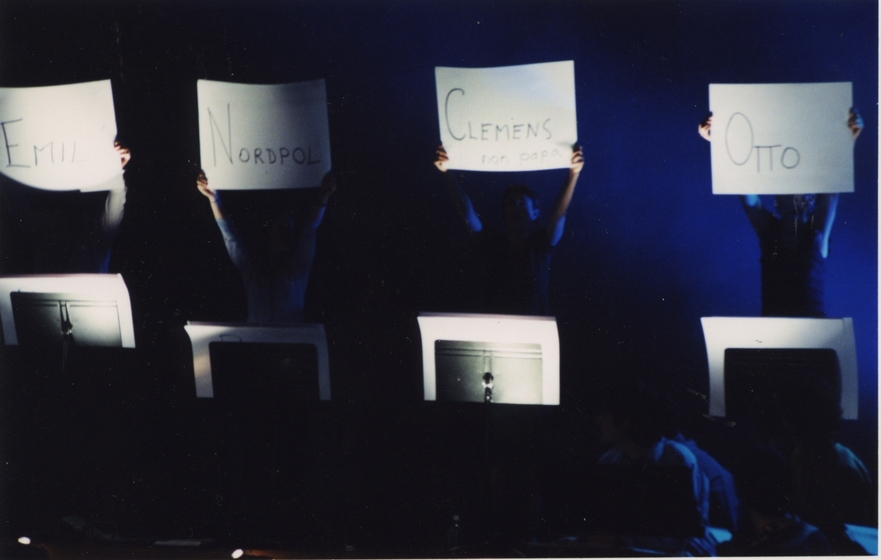 Row of four people in front of music stands, holding up white cardboard signs (L to R) read: 'Emil', 'Nordpol', 'Clemens' and 'Otto'