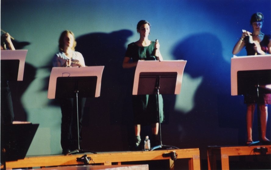 Four performers standing behind music stands, with theatre lights creating shadows behind them