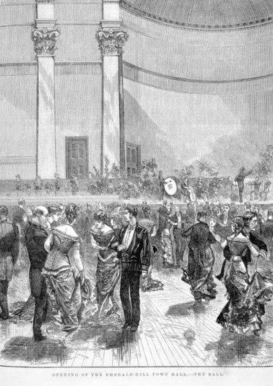 Men in suits and women in ball gowns dance in a large open ballroom, with an orchestra playing on stage behind them
