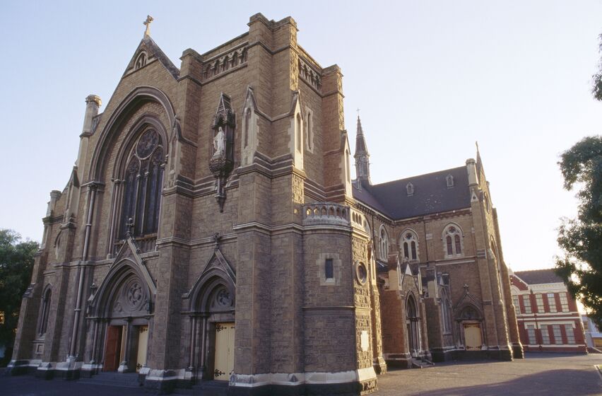 Large neo-gothic sandstone church with arched stain glass windows and wooden doors inserted into the facade