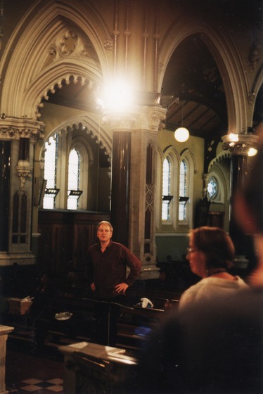 Man stands with hands on hips in a darkened church, underneath a vaulted ceiling. In the foreground, slightly out of focus stand two members of a choir.