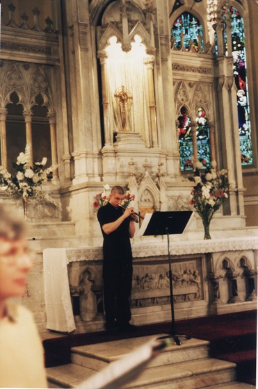 Man plays trumpet in front of a marble altar at the front of a church, flowers in vases and a golden cross adorn the altar.