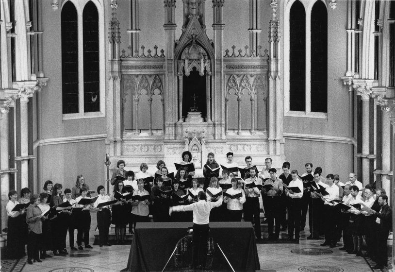 Black and White Image of choir group standing in front of an ornate altar inside a church. A conductor leads the group, while choir members hold music books and sing