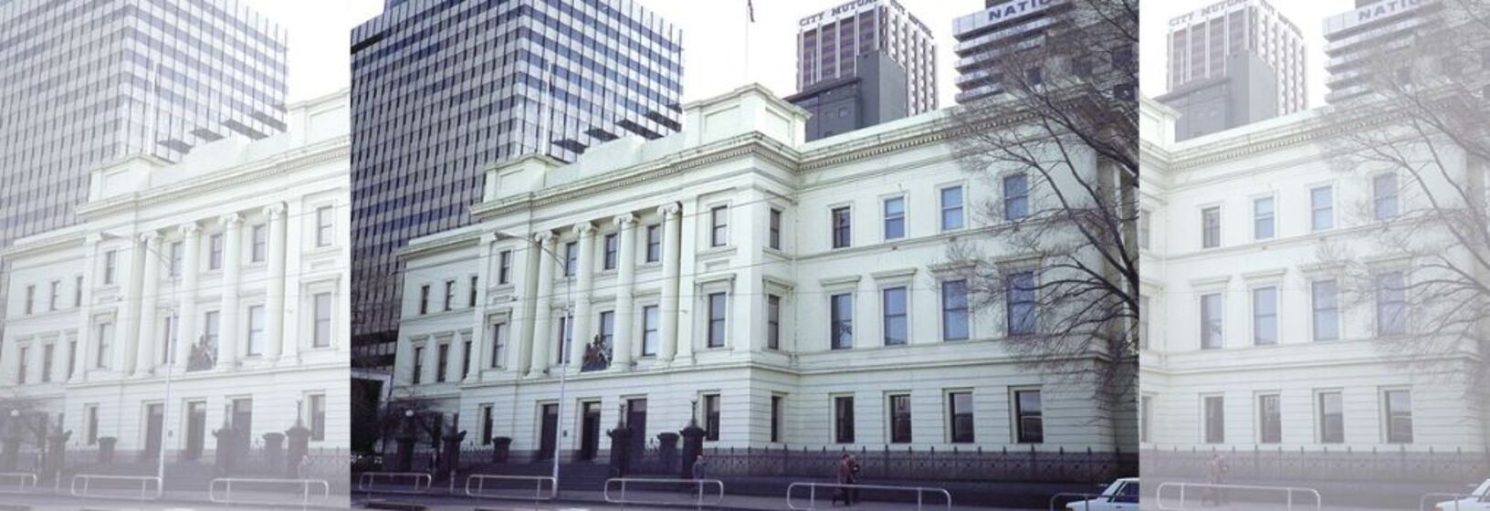 Three story white building, with multiple windows along the wide facade. City tower buildings in the background and a road with cars in the foreground.