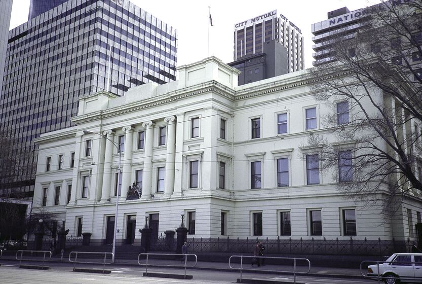 Three story white building, with multiple windows along the wide facade. City tower buildings in the background and a road with cars in the foreground.