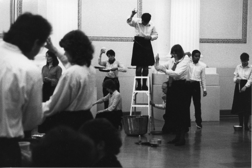 Group of performers stand around, some with their arms in the air. One woman is on a ladder in the centre of the room, looking down at the ground