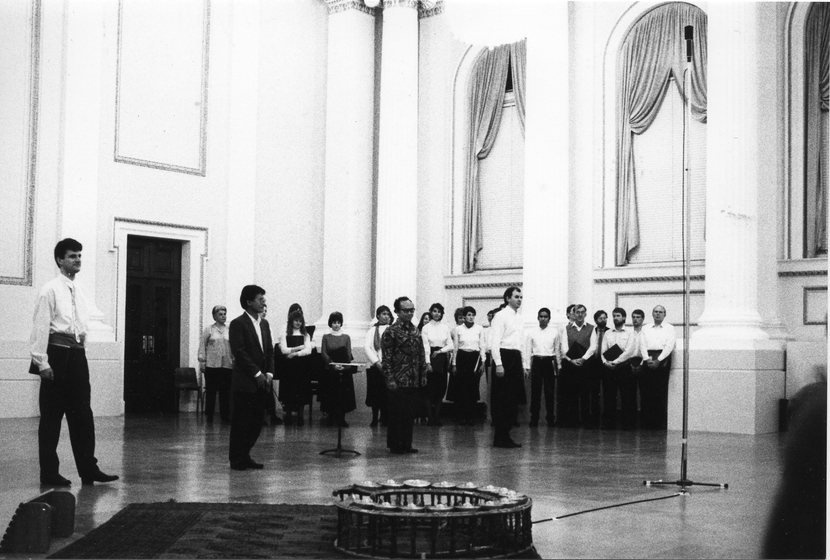 Group of performers stand in a large open space, four singers stand at the front and the rest of the group stand in front of tall columns