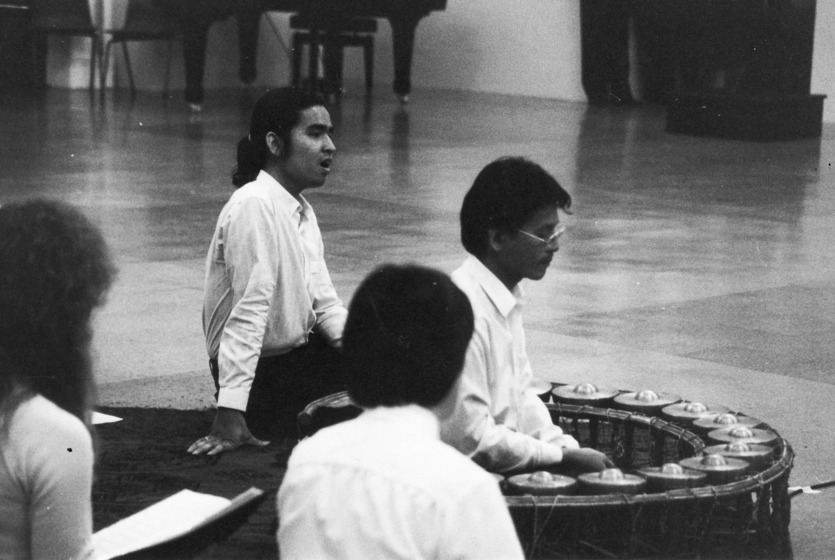 Four performers sit on the floor, one is in front of a semi circle structure with symbols on top, and another performer is singing