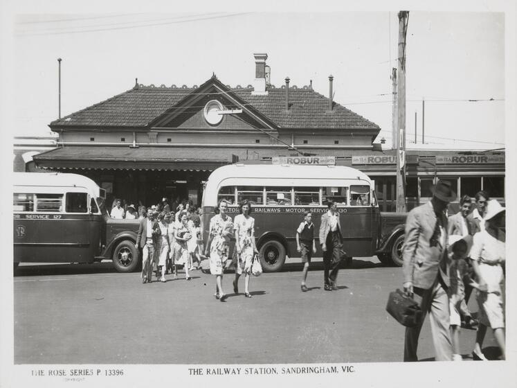 Black and white postcard with group of people exiting a  train station, older style buses are parked out the front of the station whilst people walk past them