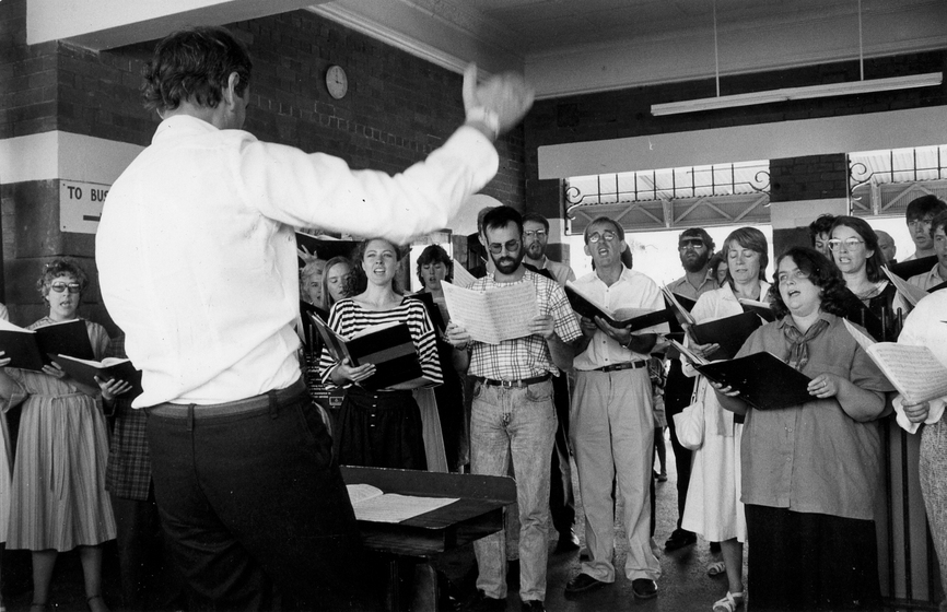 Man conducts a group of choir singers in the train station lobby, all of the singers are holding music books
