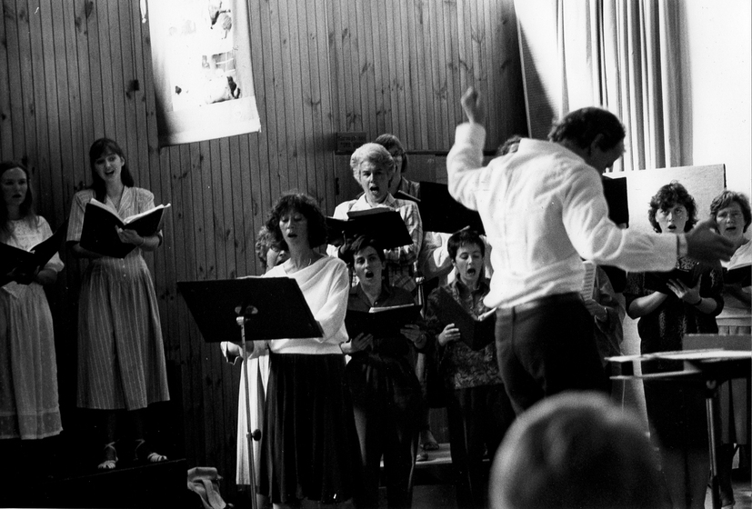 Man stands at the front, with his arm in the air, and conducts a group of 8 choral singers