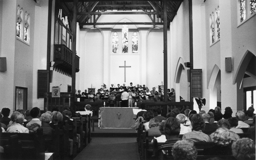 Group of people sit in a church, looking towards the altar at the front. A group of choral singer performs on the altar, all holding music books.