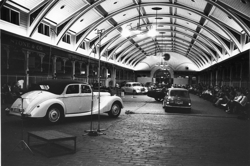 Selection of older style cars parked under a large domed roof, a crowd of seated people sits to the side and watches