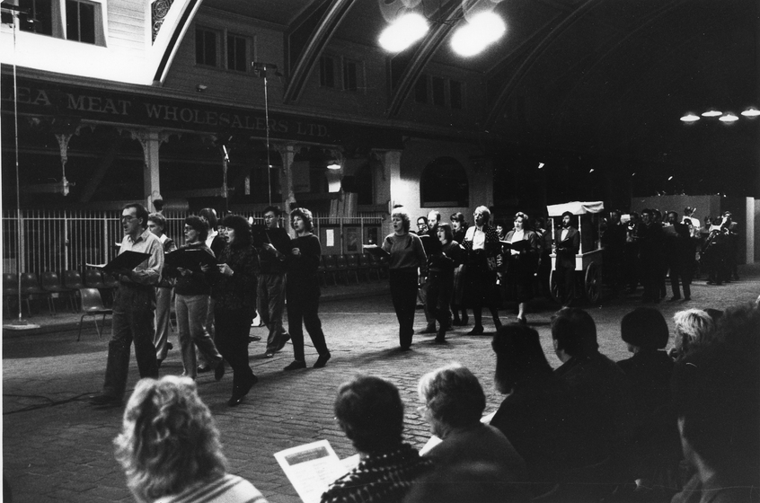A group of singers walk in a line, singing from the music books they are holding. A group of seated people watch from the sideline