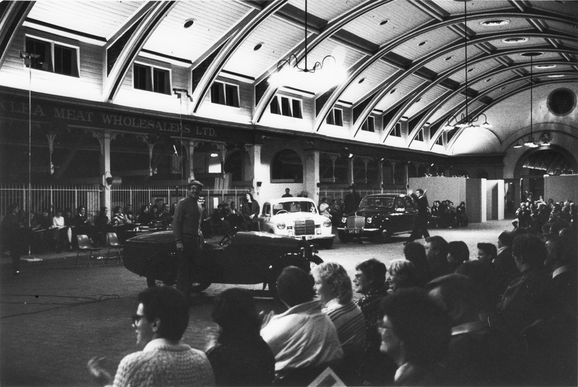 Man stands next to a small open top car while people on the sidelines watch the performers move around the cars on display