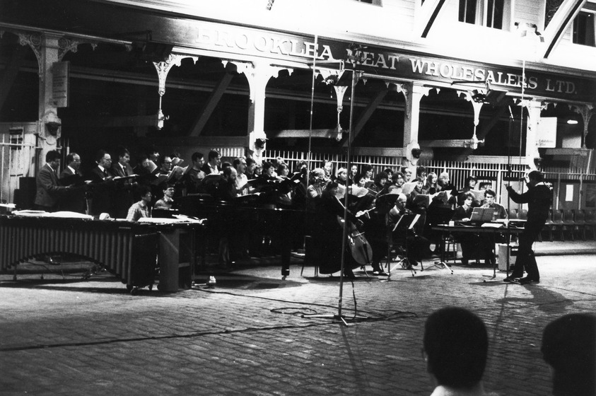 Group of standing and seated choral performers sing in an open space in an empty meat market