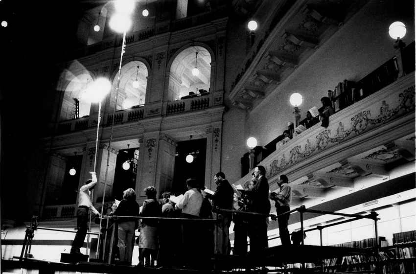A man conducts a small group of choral singers from a raised platform in the centre of a library. There are more singers standing on a balcony in the background