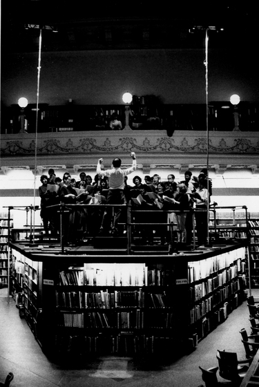 A conductor leads a group of choral singers on a raised platform that sits above a collection of library book shelves