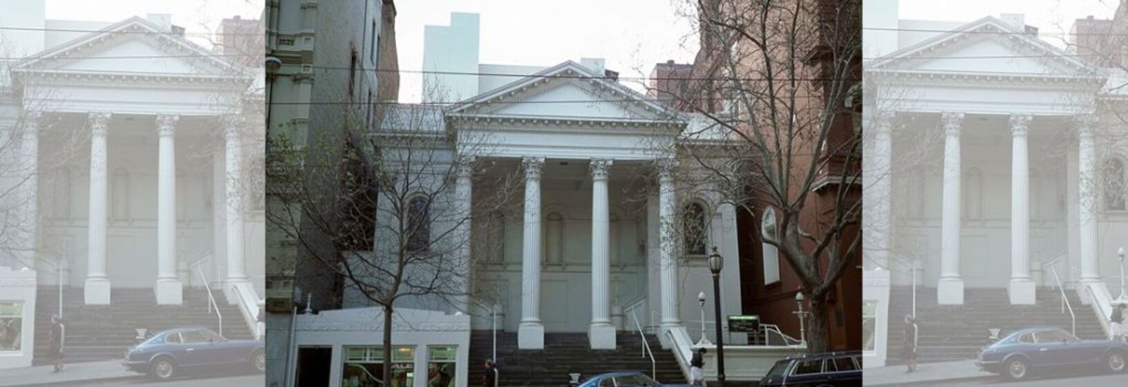 A tall white building with a columned portico at the front, trees line the street in the forefront