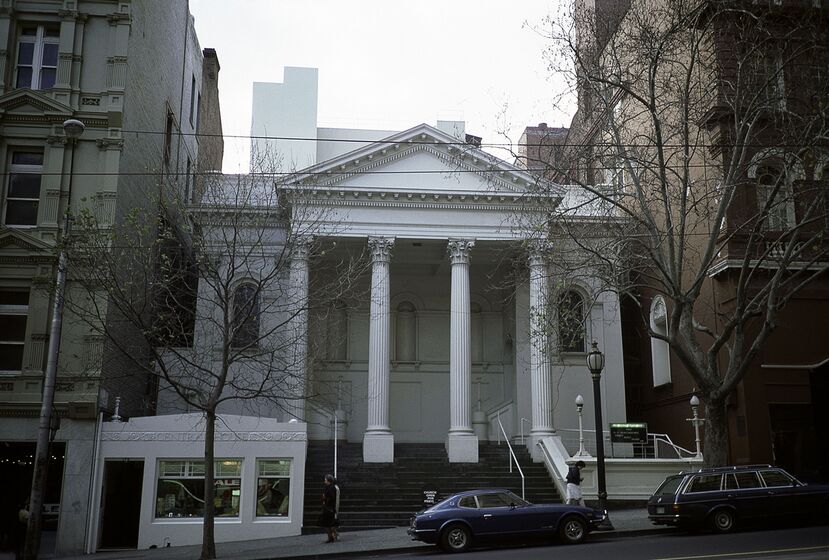 A tall white building with a columned portico at the front, trees line the street in the forefront
