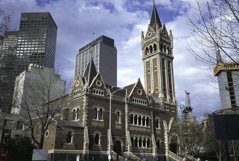 Brown brick building with tall bell tower to the right, city office towers stand in the background behind the brick building