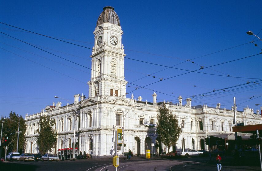 White building with tall clock tower at the front of the complex, tram tracks lie in front of the building and overhead tram wires can be seen in the foreground