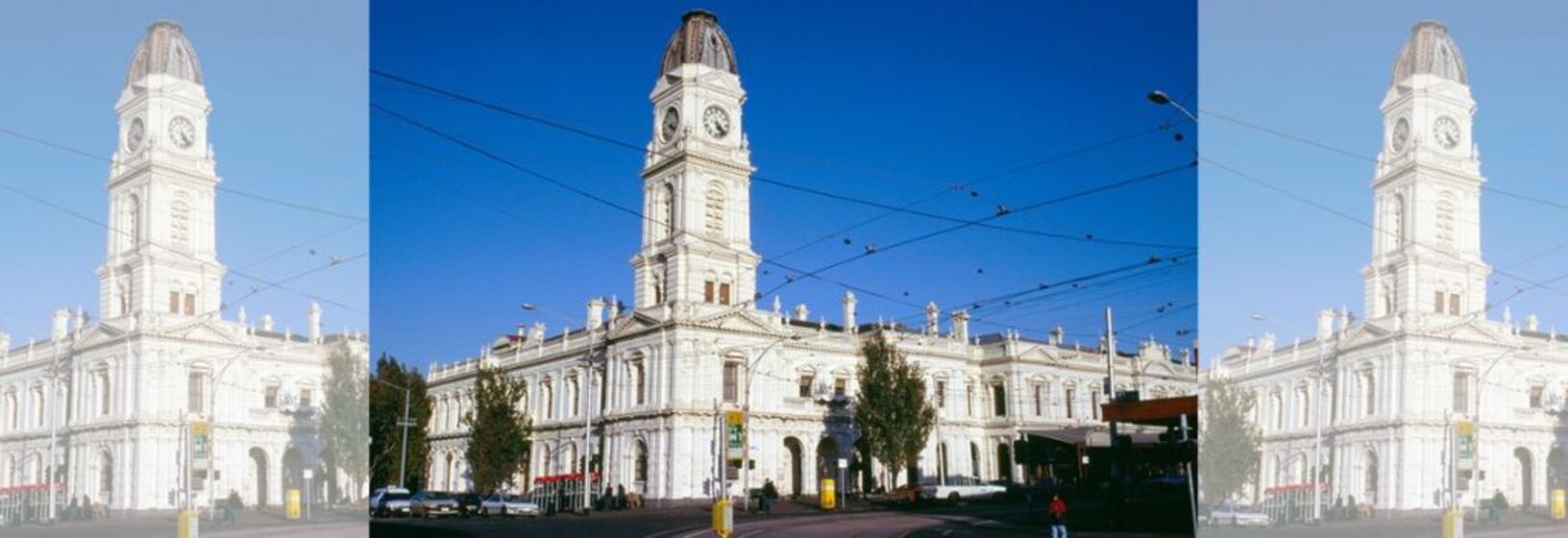 White building with tall clock tower at the front of the complex, tram tracks lie in front of the building and overhead tram wires can be seen in the foreground