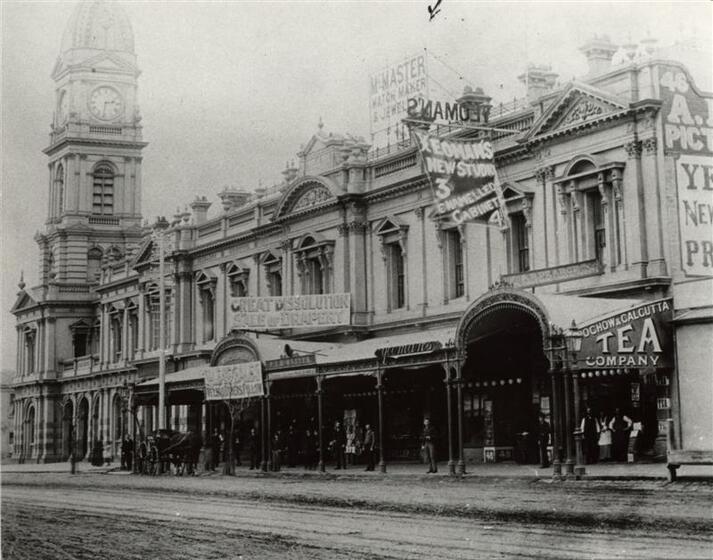 Historic Image of 19th Century building with tall clock tower at the far end. Old advertisements adorn the walls of the building