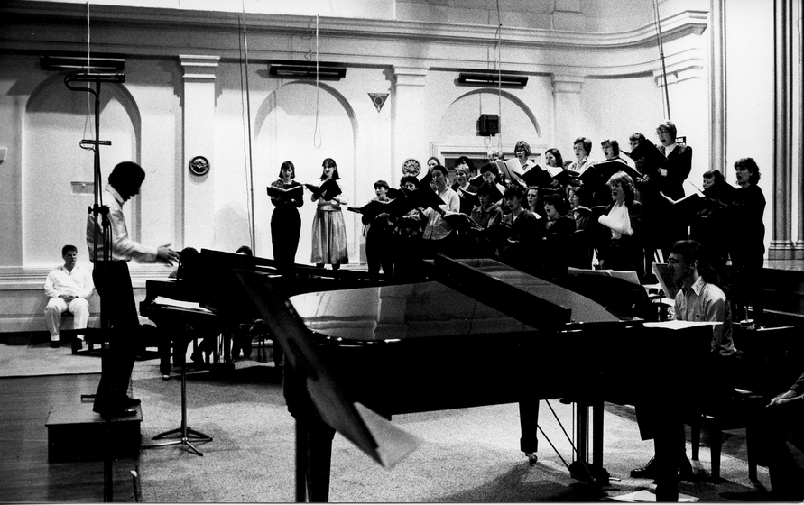 Conductor stands next to a man t a grand piano, while a group of choral singers stand behind them on an elevated platform